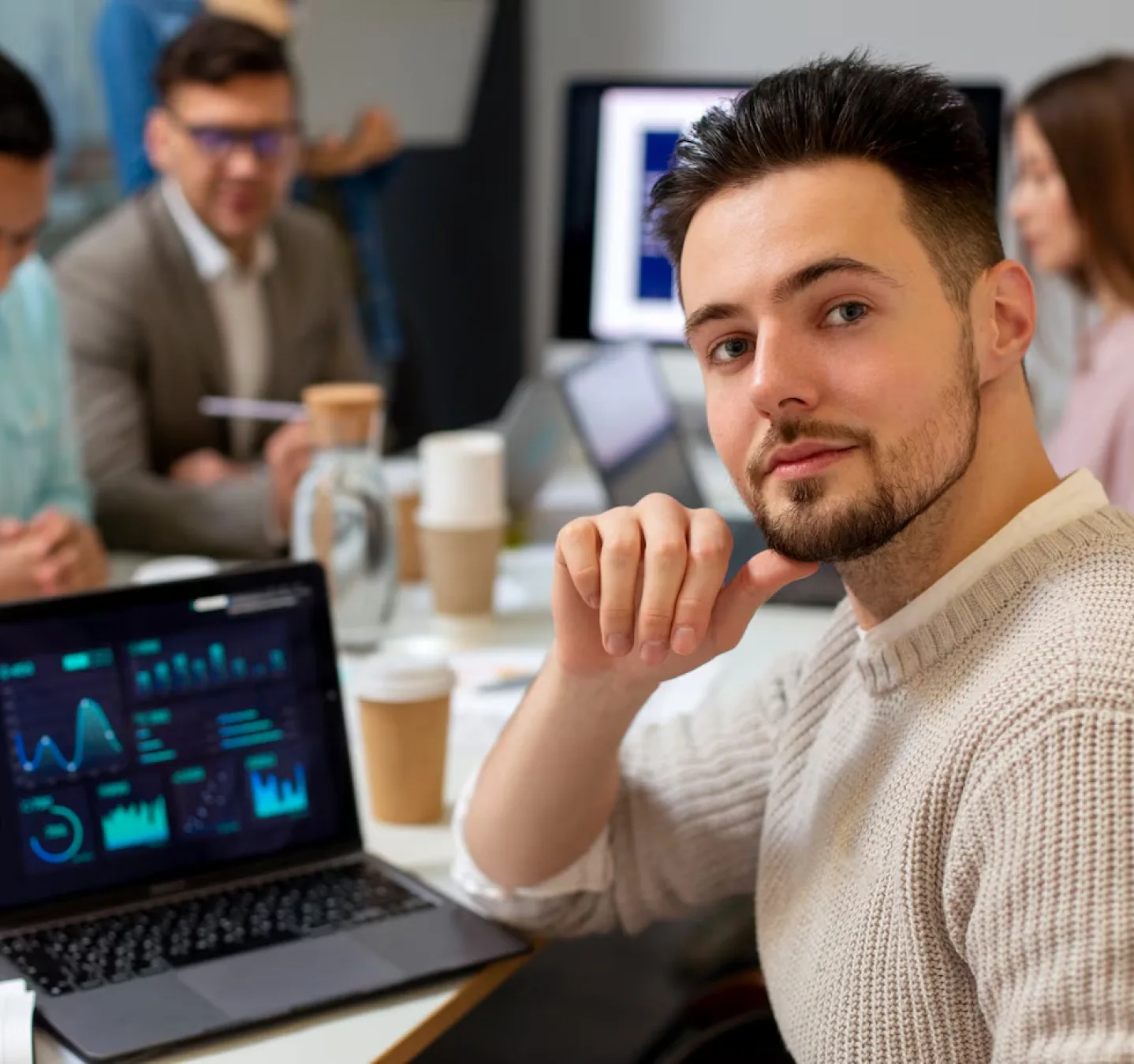 A person looks out at you with graphs of data on a laptop in the backbround